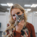 A woman with long blonde hair holds a small tabby kitten close to her face. She is wearing a rust-brown long-sleeve top, and the background shows a softly blurred indoor space with ceiling lights.